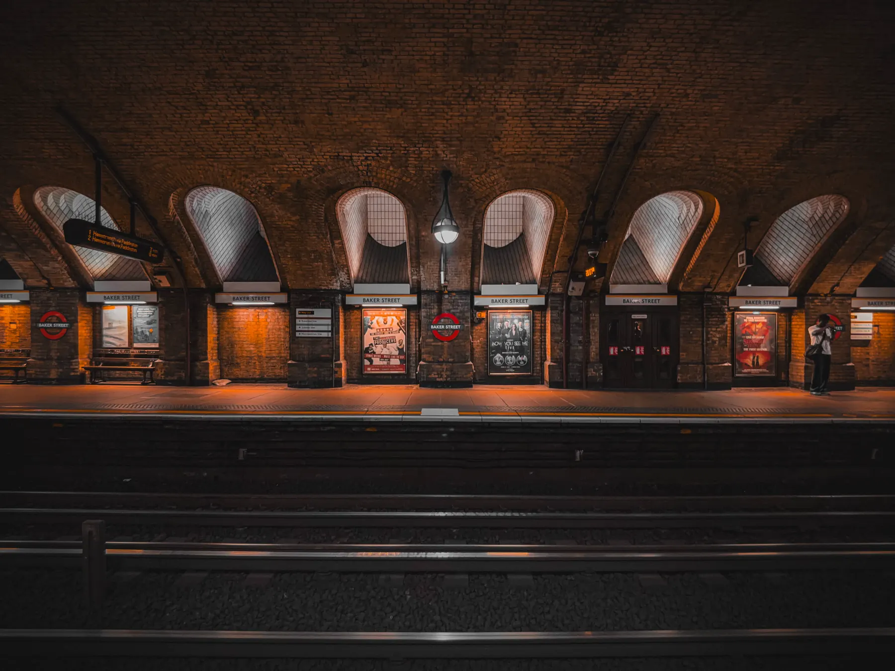 Baker Street Underground platform seen head-on from across the tracks: a row of brick arches glazed with fanlight windows, warm sodium light, a lone photographer at the far right.